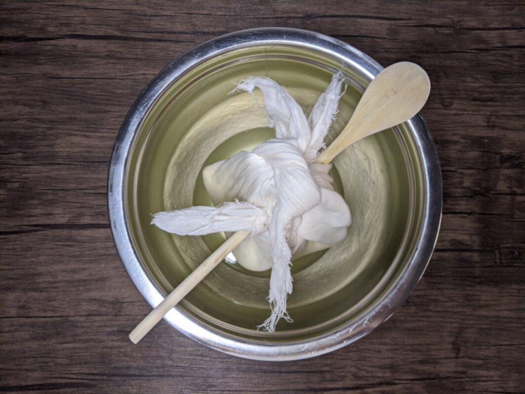 Jogurt zawinięty w płótno, podwieszony na drewnianej łyżce nad miską, odcieka z serwatki.